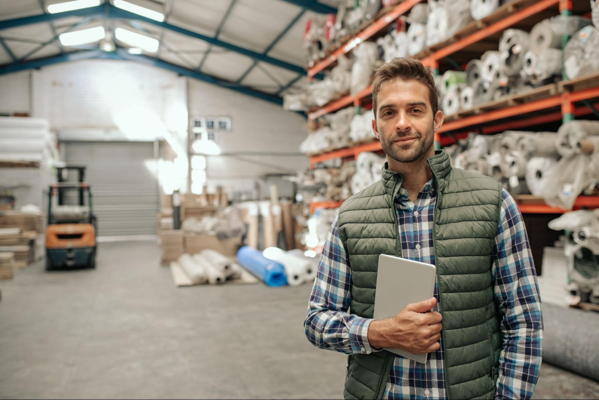 Man in a plaid shirt and green vest holding a tablet, standing in a warehouse filled with rolls of fabric and materials on shelves, with a forklift in the background.